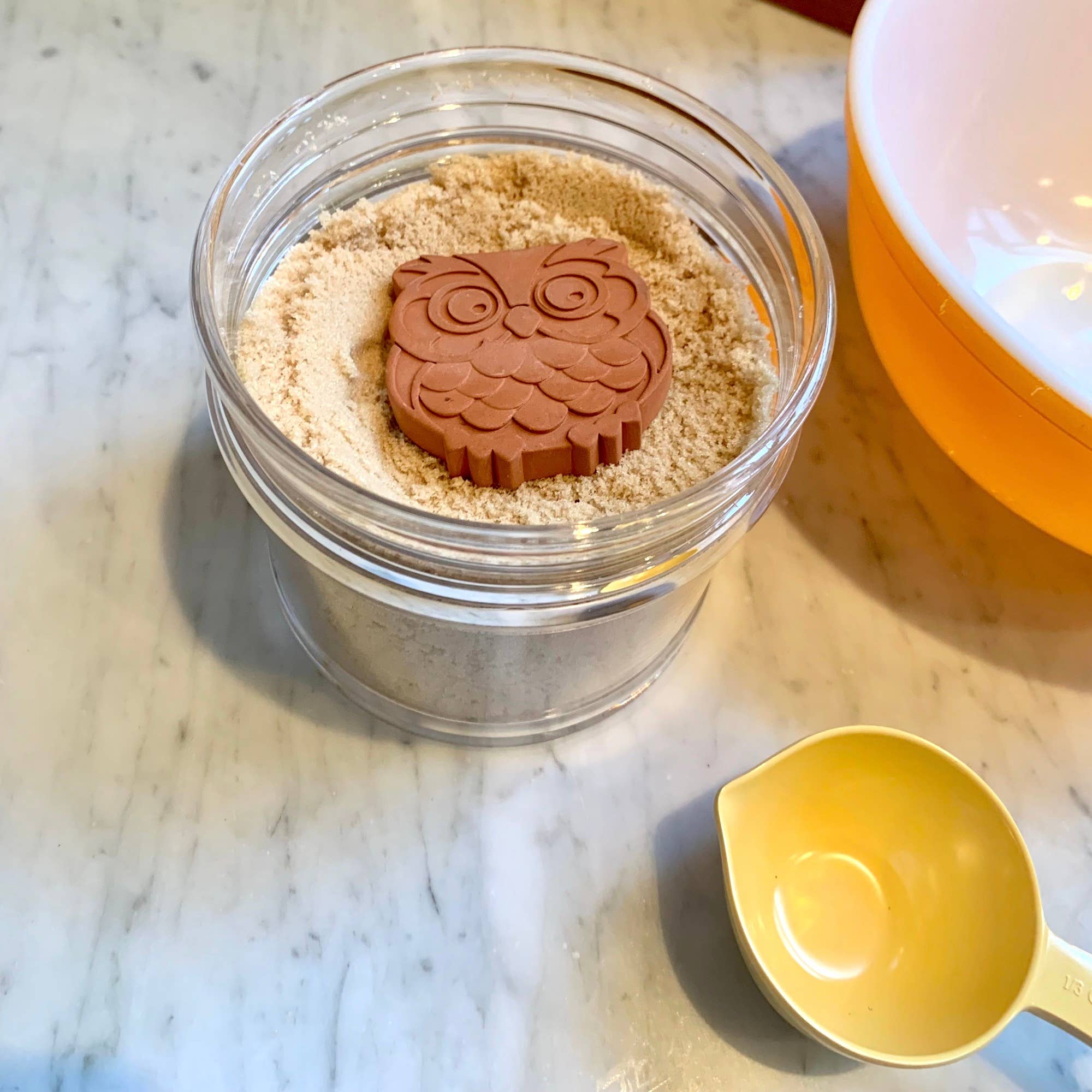 Jar of sand with an owl-shaped mold on a marble surface next to a yellow scoop.