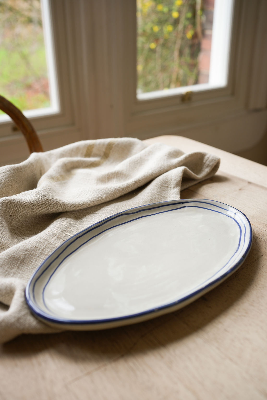 oval-pottery-plate-with-blue-stripe-border-on-wooden-table-with-linen-teatowel-windows-in-back-ground,