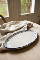 oval-pottery-plate-with-blue-stripe-border-on-wooden-table-with-linen-teatowel-windows-in-back-ground,