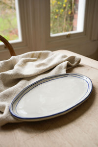 oval-pottery-plate-with-blue-stripe-border-on-wooden-table-with-linen-teatowel-windows-in-back-ground,