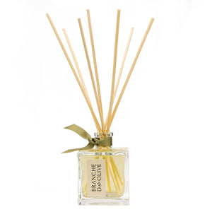 A clear glass room diffuser bottle with a label, containing several light brown reeds and a green decorative ribbon at the top.