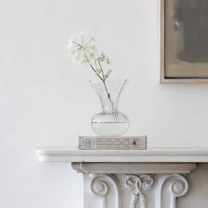 A clear glass vase with a flared neck and rounded base sits on a white mantelpiece, holding a single white flower. A vintage book rests beneath the vase.