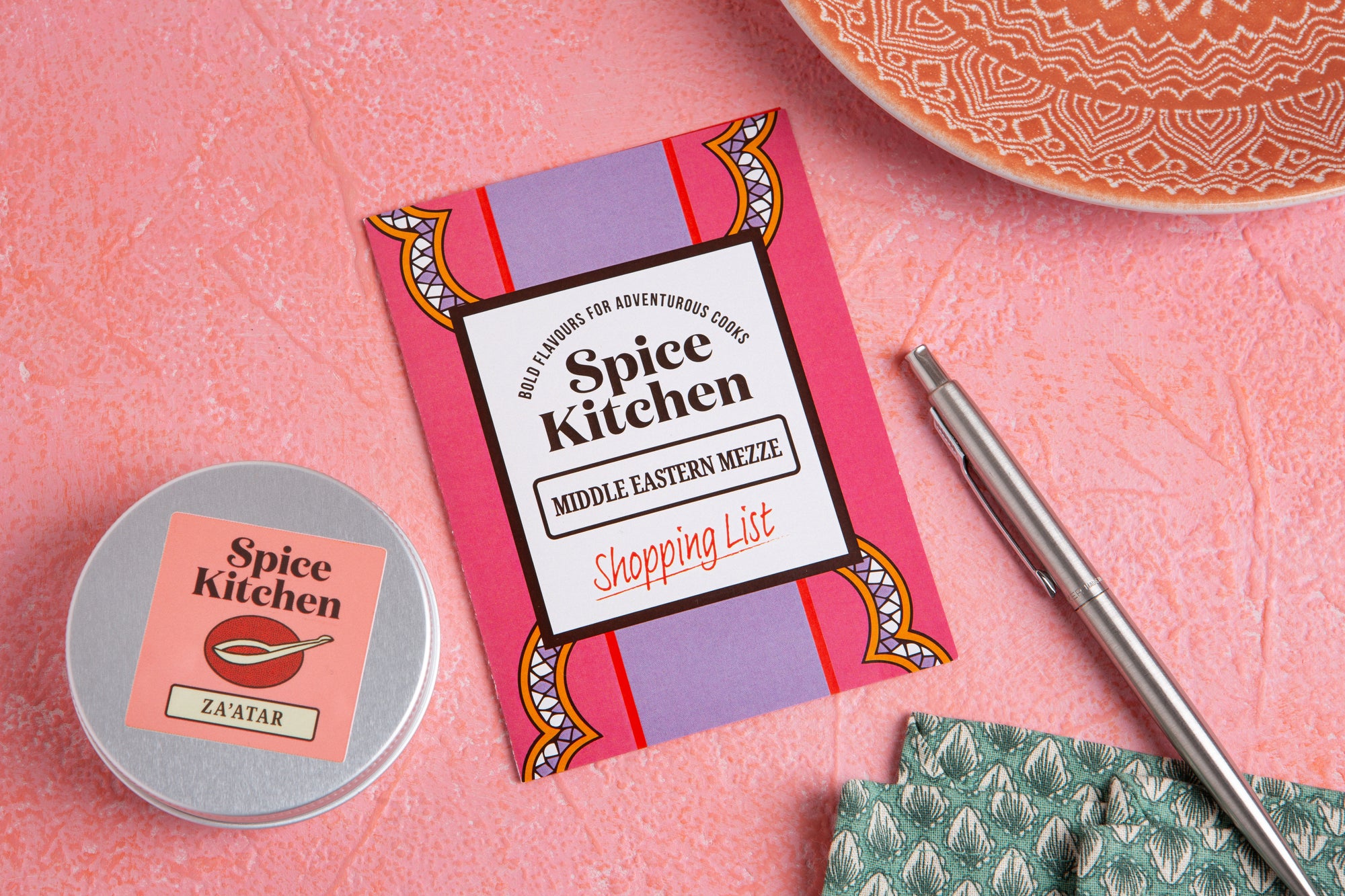 Spice Kitchen product packaging on a pink surface with a pen and container.