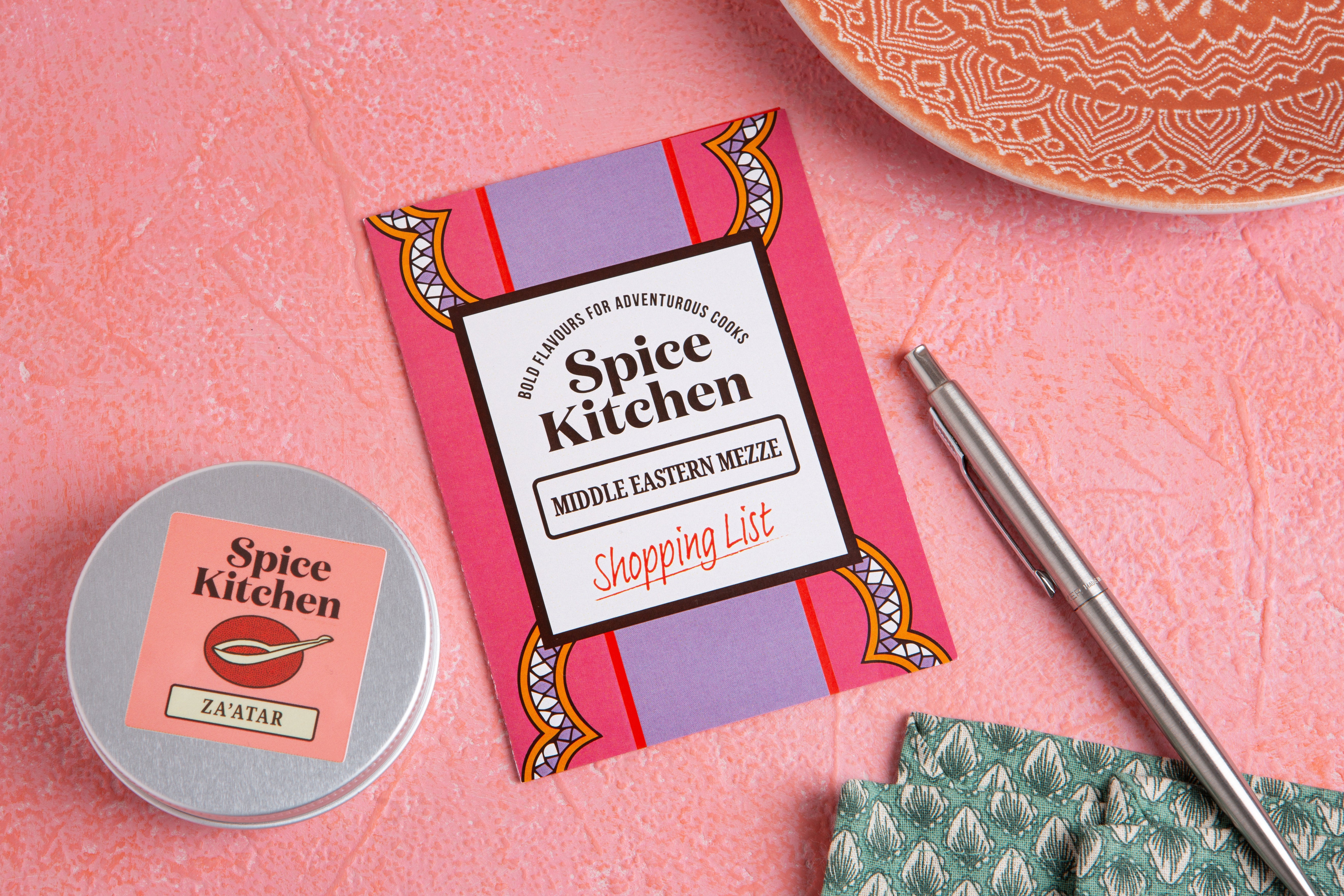 Spice Kitchen product packaging on a pink surface with a pen and container.